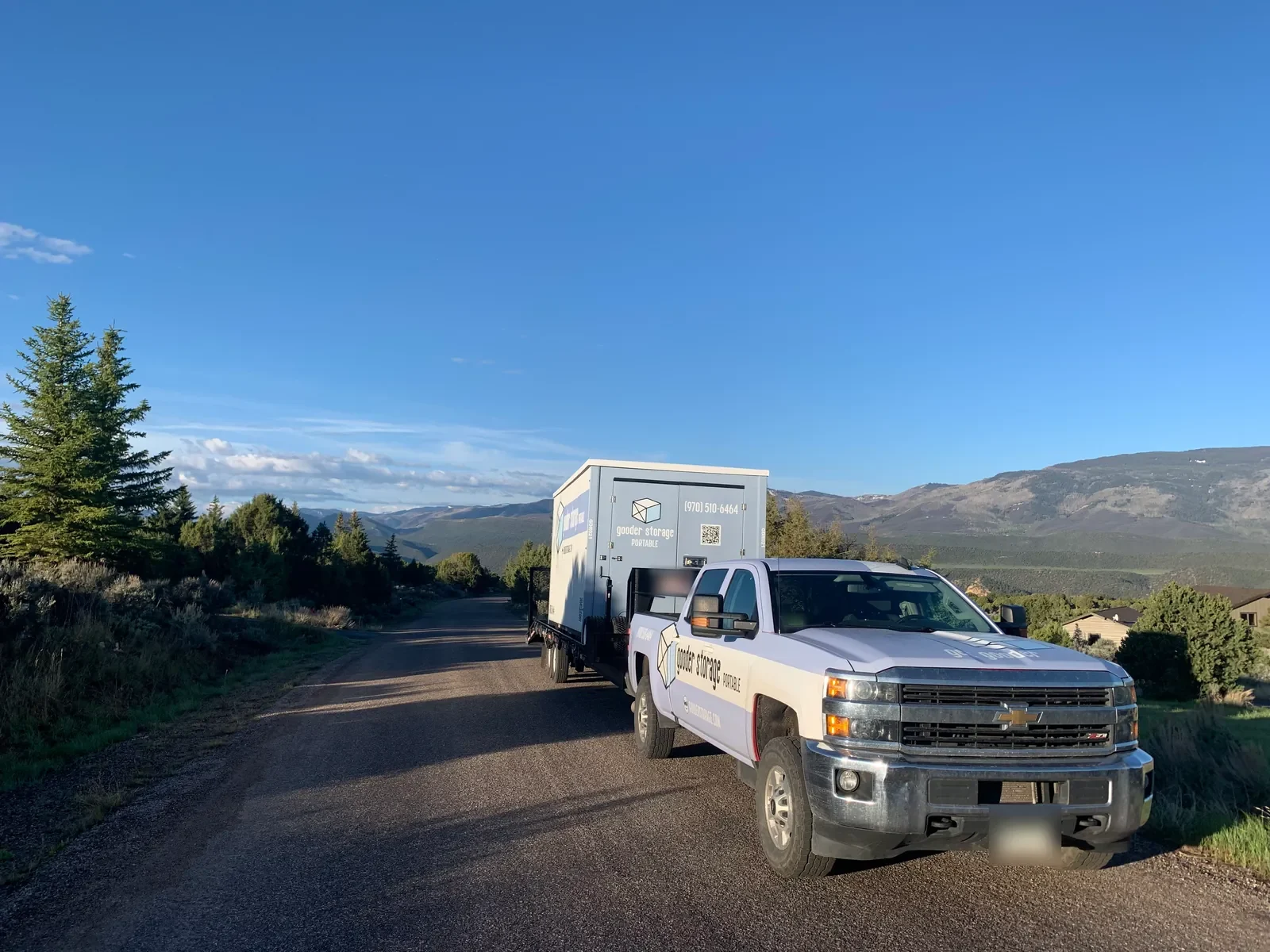 Gooder truck towing a Gooder box on a trailer down a quiet gravel road, with mountains and blue sky behind